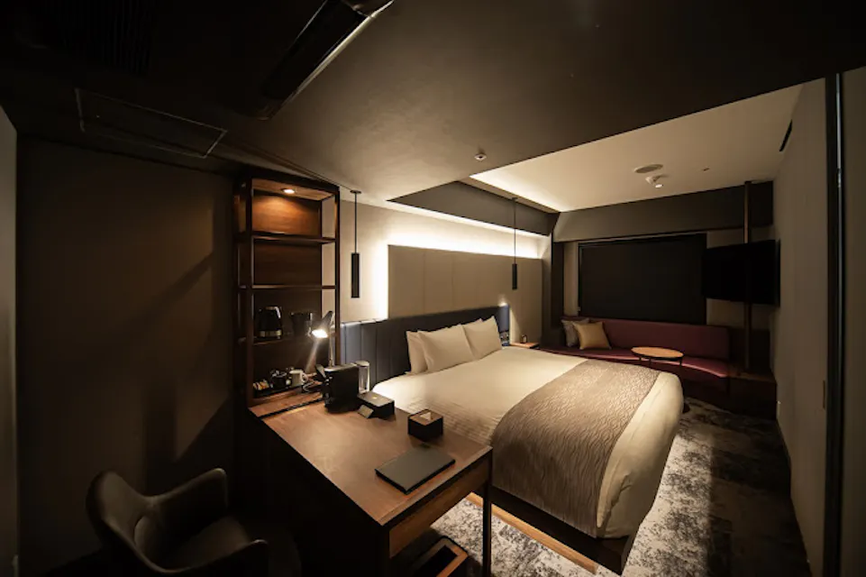 A modern hotel room features a cozy double bed with a light brown blanket and white pillows. To the left is a wooden desk with a lamp and chair, while to the right sits a maroon sofa under a large window, with a wall-mounted TV nearby. Warm lighting creates a comfortable ambiance.
