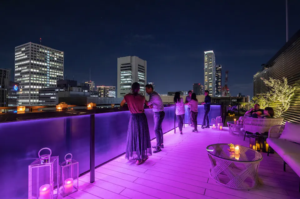 A rooftop bar at night with patrons enjoying the city skyline. The setting features purple lighting, modern decor, candles on tables, and city buildings illuminated in the background. People are standing and conversing near the railing, appreciating the night view.