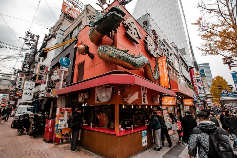 日本の賑やかな都市エリアのストリートビュー。鮮やかな建物には赤い外観に大きな緑のドラゴン像が飾られており、角には忙しい屋台があります。人々は並んで待っており、さまざまな看板や電線、背景に高い建物に囲まれています。