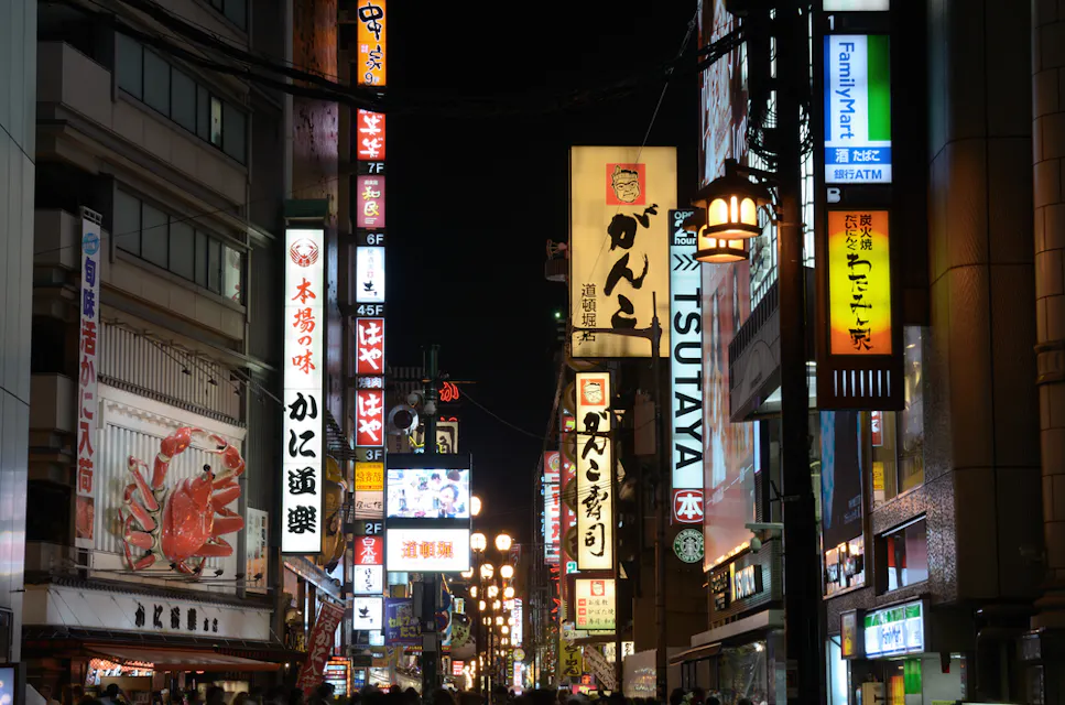 日本の夜の賑やかな街の景色。様々な言語（日本語を含む）の明るく照らされた店舗の看板が並ぶ。看板は、レストラン、ショップ、その他のビジネスを宣伝している。ネオンの光とカラフルなディスプレイが活気ある都市の雰囲気を支配している。