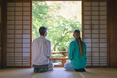 A man and a woman kneel on a tatami floor, facing an open shoji screen and looking out at a lush green garden, with soft natural light streaming in.