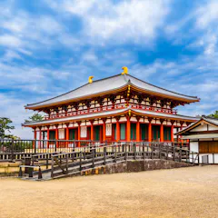 Kofuku-ji Temple A traditional Japanese temple with a red and white facade, ornate roof, and golden ornaments stands under a partly cloudy sky, surrounded by a courtyard and trees.