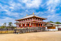A traditional Japanese temple with a red and white facade, ornate roof, and golden ornaments stands under a partly cloudy sky, surrounded by a courtyard and trees.
