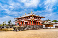 A traditional Japanese temple with a red and white facade, ornate roof, and golden ornaments stands under a partly cloudy sky, surrounded by a courtyard and trees.