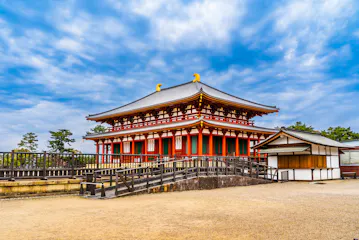 A traditional Japanese temple with a red and white facade, ornate roof, and golden ornaments stands under a partly cloudy sky, surrounded by a courtyard and trees.