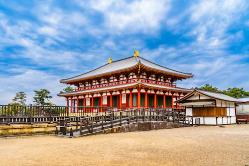 A traditional Japanese temple with a red and white facade, ornate roof, and golden ornaments stands under a partly cloudy sky, surrounded by a courtyard and trees.