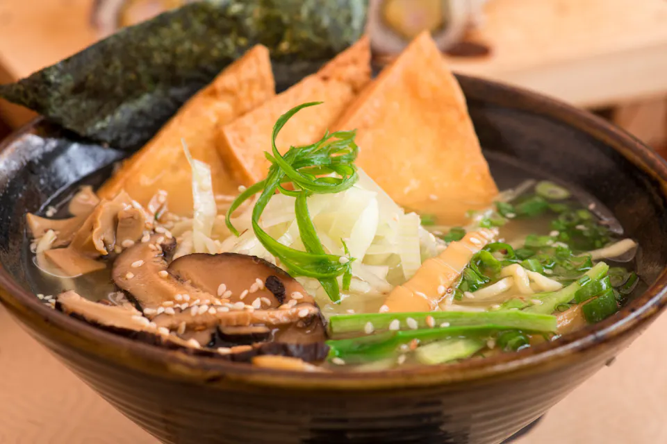 A bowl of ramen topped with sliced mushrooms, tofu triangles, seaweed, green onions, and sesame seeds. The broth looks savory and is garnished with sliced leeks, providing a fresh and appetizing appearance.