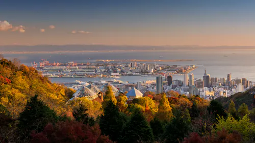 A panoramic view of Kobe, Japan at sunset, showing city buildings, a waterfront port area, and distant mountains, with autumn-colored trees in the foreground.