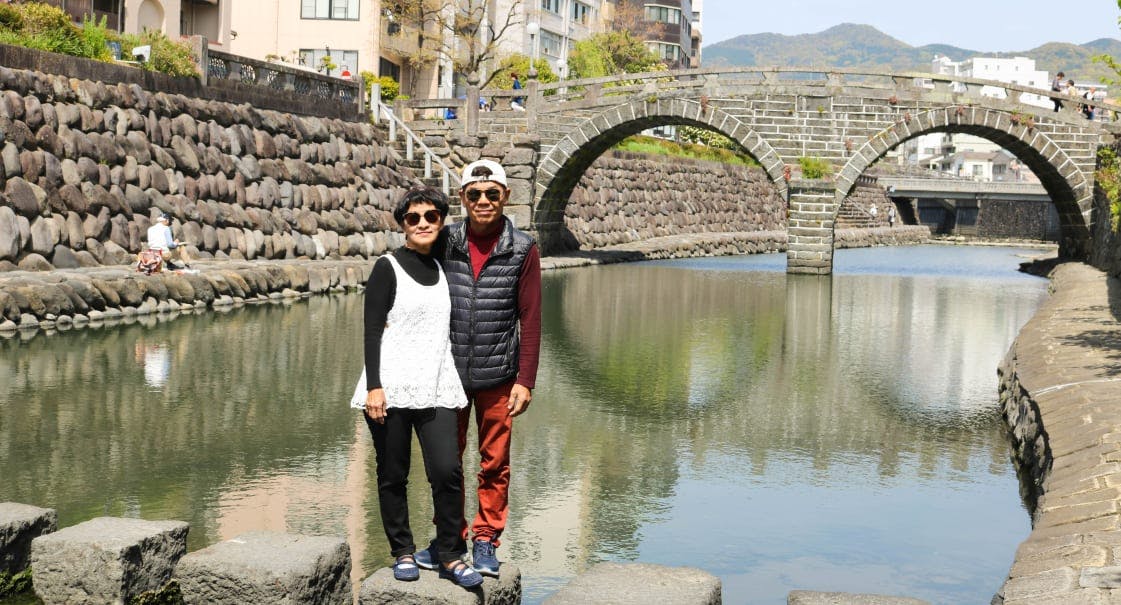Two people stand smiling beside a stone-lined canal, with a historic arched stone bridge and buildings in the background. The sky is clear and the water reflects the bridge.