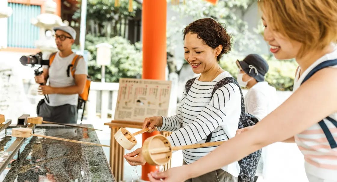 Two women smile while using wooden ladles at a water basin in a Japanese shrine, performing a purification ritual. A man with a camera stands in the background, observing the scene.