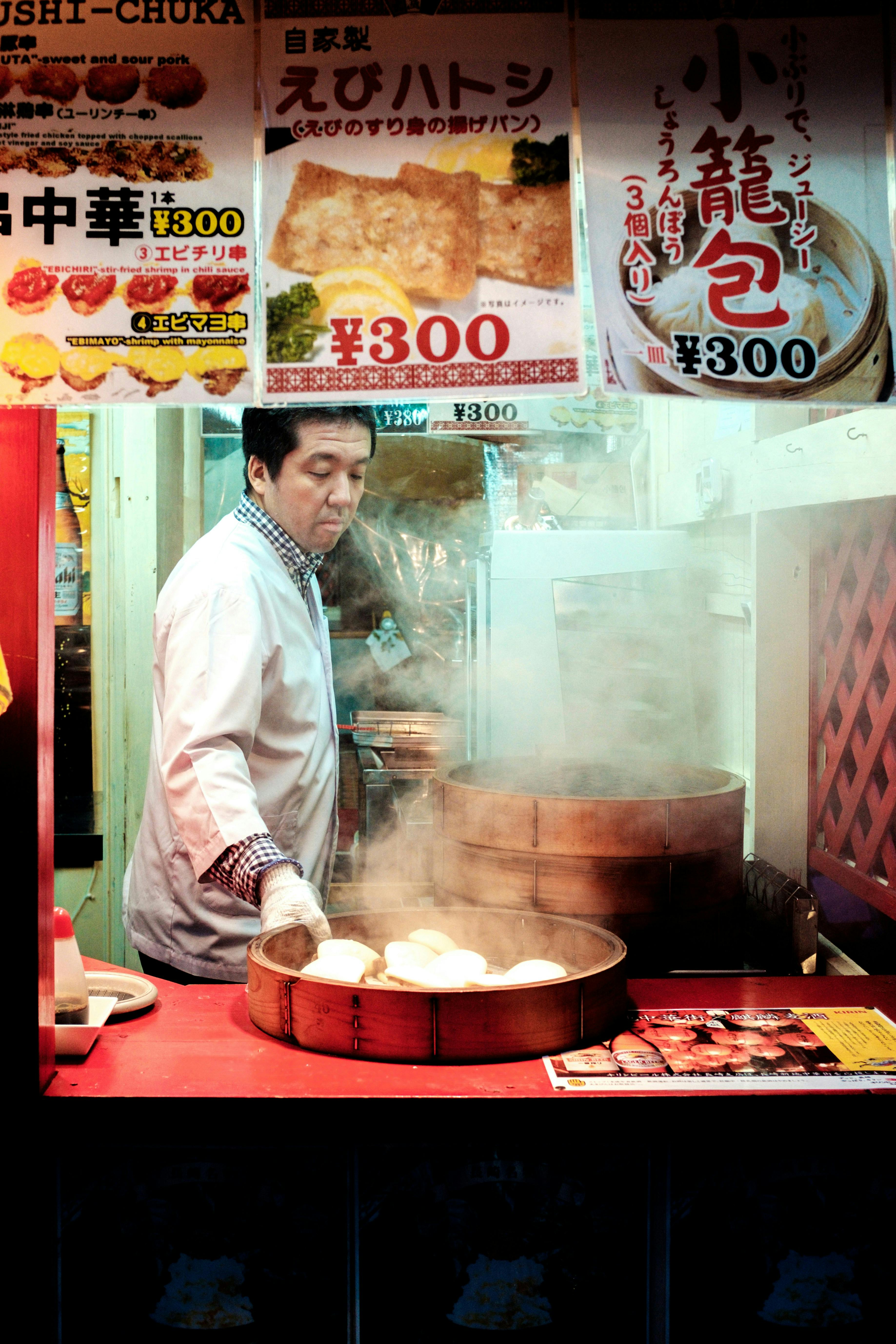 A vendor steams buns in a bamboo basket at a food stall, surrounded by signs with Japanese writing and prices. Steam rises from the buns, and colorful advertisements hang above the stall.