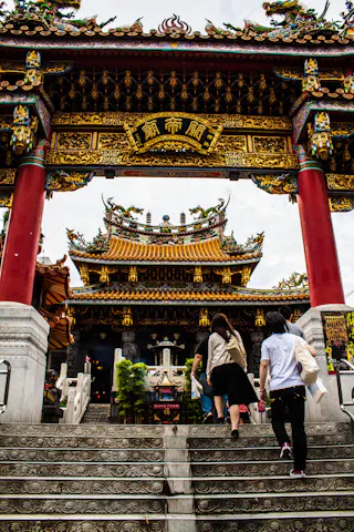 People walk up stone steps toward a colorful, ornate traditional Chinese temple gate with golden details and red pillars, leading to a richly decorated temple building under a cloudy sky.