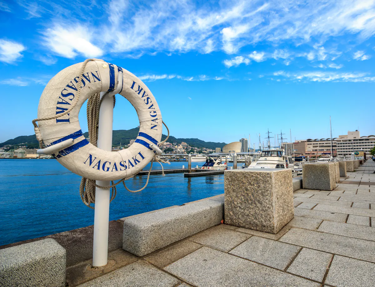 Dejima A lifebuoy labeled "Nagasaki" is attached to a post on a waterfront promenade, overlooking a marina with boats, buildings, and distant green hills under a partly cloudy blue sky.