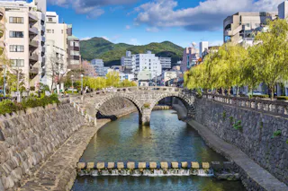A stone arch bridge spans a calm river lined with stone walls and walkways, surrounded by modern buildings and lush green trees, with mountains in the background under a blue sky with clouds.