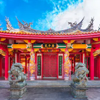 Kōshi-byō A vibrant traditional Chinese temple with ornate roof decorations, red pillars, intricate carvings, and two stone lion statues at the entrance under a blue sky.