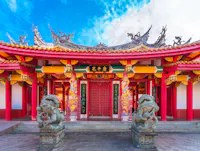 A vibrant traditional Chinese temple with ornate roof decorations, red pillars, intricate carvings, and two stone lion statues at the entrance under a blue sky.
