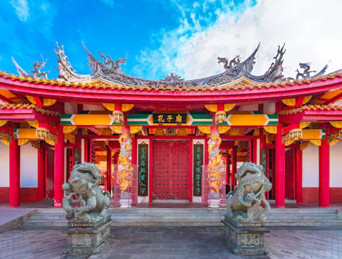 A vibrant traditional Chinese temple with ornate roof decorations, red pillars, intricate carvings, and two stone lion statues at the entrance under a blue sky.