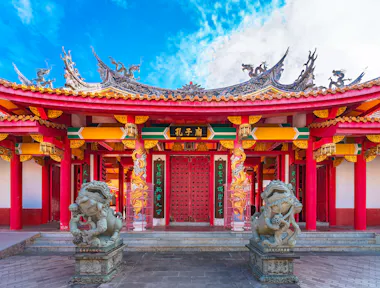 A vibrant traditional Chinese temple with ornate roof decorations, red pillars, intricate carvings, and two stone lion statues at the entrance under a blue sky.