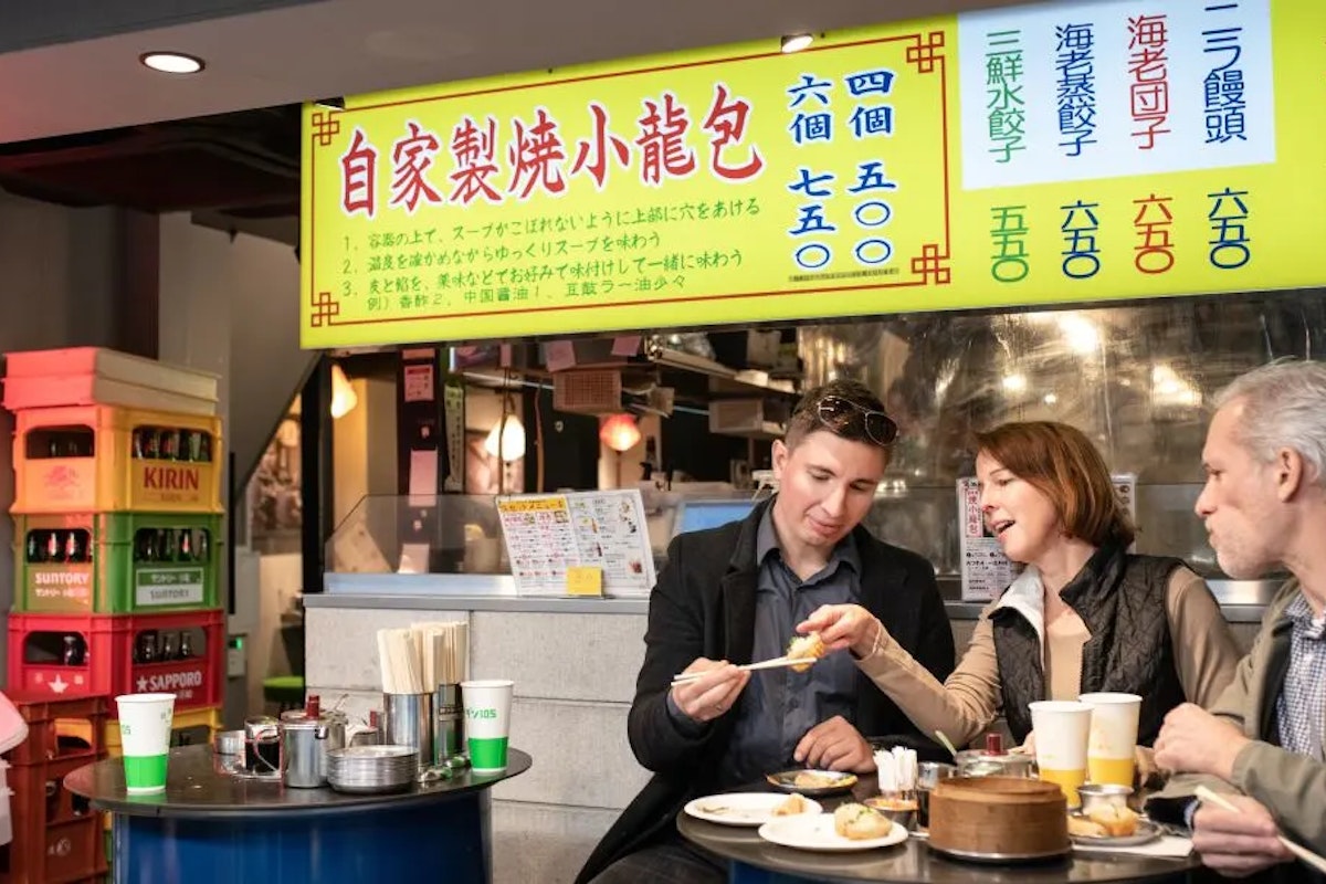 Tokyo Food Tour Three people sit at a table in a restaurant, eating dumplings. They are sharing conversation and food. Bright yellow signs with Chinese characters and colorful labels hang in the background. A fridge with drinks is visible on the left.