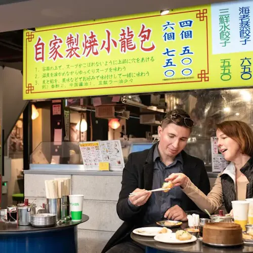 Tokyo Food Tour Three people sit at a table in a restaurant, eating dumplings. They are sharing conversation and food. Bright yellow signs with Chinese characters and colorful labels hang in the background. A fridge with drinks is visible on the left.