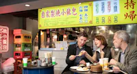 Three people sit at a table in a restaurant, eating dumplings. They are sharing conversation and food. Bright yellow signs with Chinese characters and colorful labels hang in the background. A fridge with drinks is visible on the left.