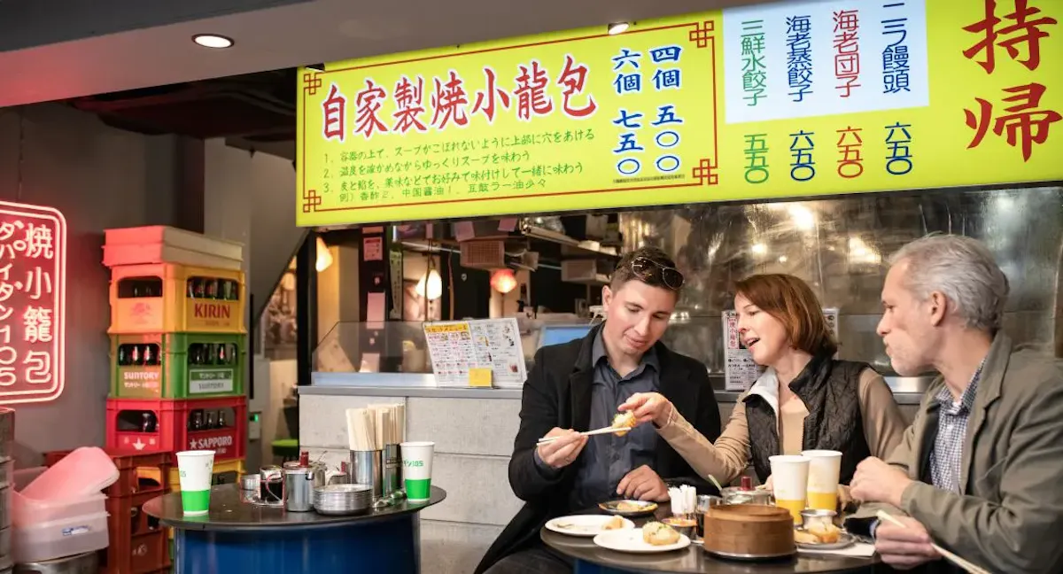 Three people sit at a table in a restaurant, eating dumplings. They are sharing conversation and food. Bright yellow signs with Chinese characters and colorful labels hang in the background. A fridge with drinks is visible on the left.