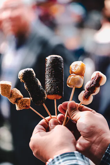 A person holds five skewers of various Japanese street foods, including rice cakes coated in seaweed and sweet sauces, with a blurred crowd in the background.