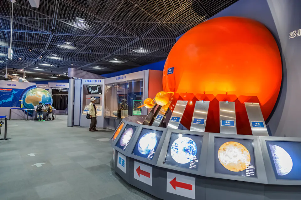 科学博物館の展示で、大きく照らされたオレンジ色の球体が太陽を表しています。様々な惑星モデルがその下に展示されています。情報掲示板と矢印標識があり、訪問者が展示物やインタラクティブなディスプレイを観察している様子があります。