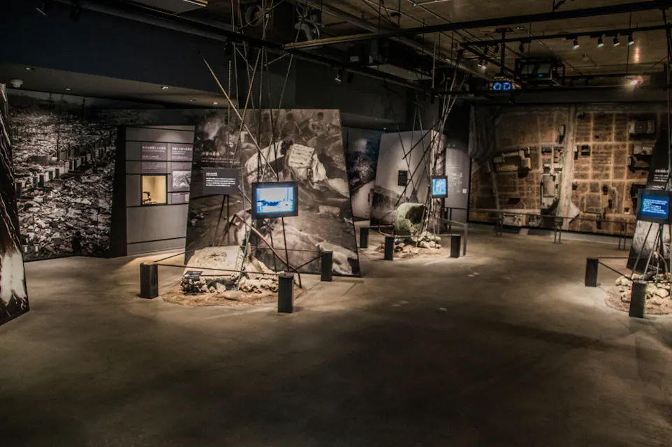 さまざまなマルチメディアディスプレイと遺物を展示する博物館の展示。部屋は薄暗く、スポットライトが異なるセクションを照らしています。大きな白黒写真が壁の一部を覆い、訪問者に情報を提供する小さなスクリーンがあります。