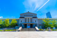 A large, symmetrical building with a triangular roof and multiple windows, set against a bright blue sky. The structure is surrounded by greenery on either side, with people and stairs leading up to the entrance. A tall skyscraper is visible in the background.
