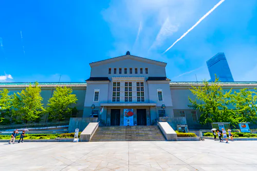 A large, symmetrical building with a triangular roof and multiple windows, set against a bright blue sky. The structure is surrounded by greenery on either side, with people and stairs leading up to the entrance. A tall skyscraper is visible in the background.