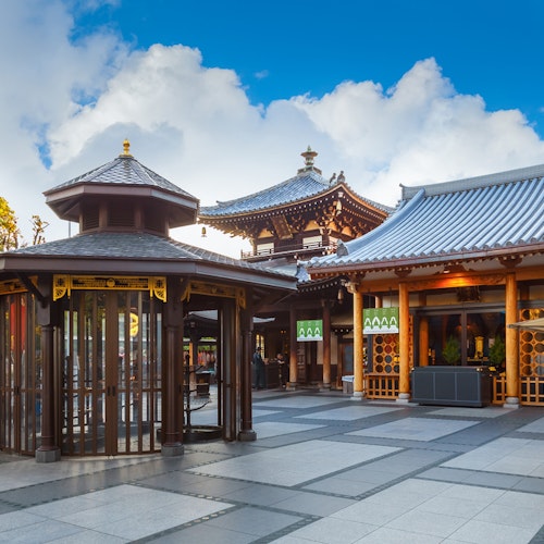 Isshinji Temple in Osaka, Japan A well-maintained Buddhist temple with traditional architecture featuring curved rooflines and wooden accents. In the foreground is a gazebo-like structure with glass windows. The sky above is bright with scattered clouds. Trees are visible in the background.