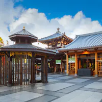 Isshinji Temple in Osaka, Japan A well-maintained Buddhist temple with traditional architecture featuring curved rooflines and wooden accents. In the foreground is a gazebo-like structure with glass windows. The sky above is bright with scattered clouds. Trees are visible in the background.