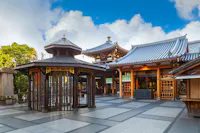 A well-maintained Buddhist temple with traditional architecture featuring curved rooflines and wooden accents. In the foreground is a gazebo-like structure with glass windows. The sky above is bright with scattered clouds. Trees are visible in the background.