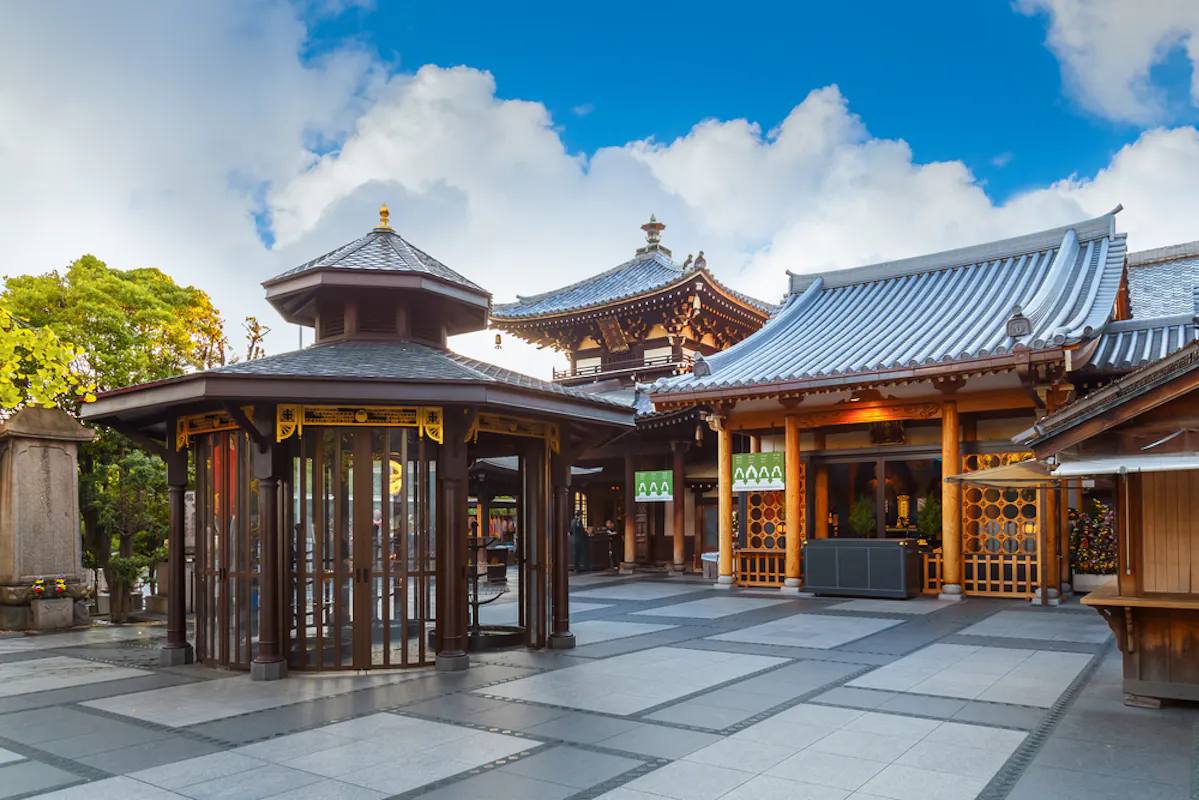A well-maintained Buddhist temple with traditional architecture featuring curved rooflines and wooden accents. In the foreground is a gazebo-like structure with glass windows. The sky above is bright with scattered clouds. Trees are visible in the background.