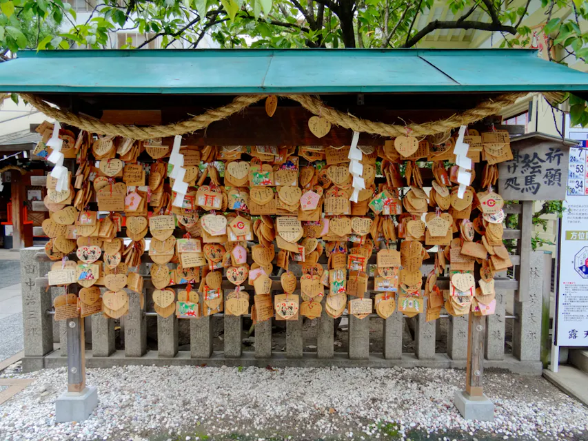 Picture of many Ema's (votive plaques) hanging at the Tsuyunoten Shrine in Osaka A wooden structure with a green roof in a Japanese shrine is adorned with numerous wooden prayer plaques, known as ema. The plaques are of various shapes and sizes, including hearts, and are inscribed with handwritten prayers and wishes. A tree stands behind it.