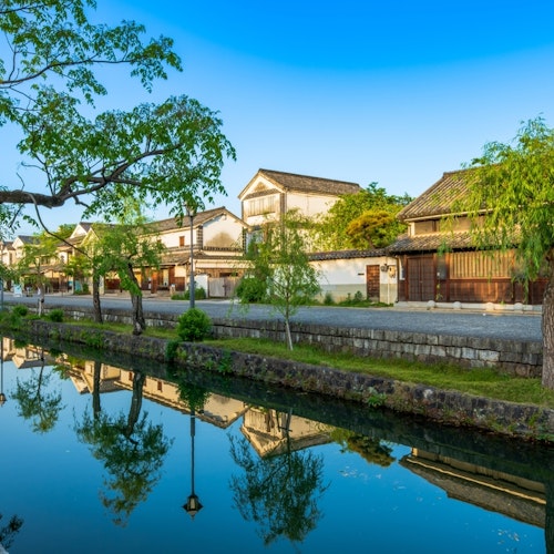The old townscape of Kurashiki City, Okayama Prefecture A serene canal lined with lush green trees reflects traditional Japanese buildings under a clear blue sky. The scene is tranquil, with historic architecture and vibrant greenery mirrored in the still water, evoking a sense of peace and timeless beauty.