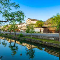 The old townscape of Kurashiki City, Okayama Prefecture A serene canal lined with lush green trees reflects traditional Japanese buildings under a clear blue sky. The scene is tranquil, with historic architecture and vibrant greenery mirrored in the still water, evoking a sense of peace and timeless beauty.