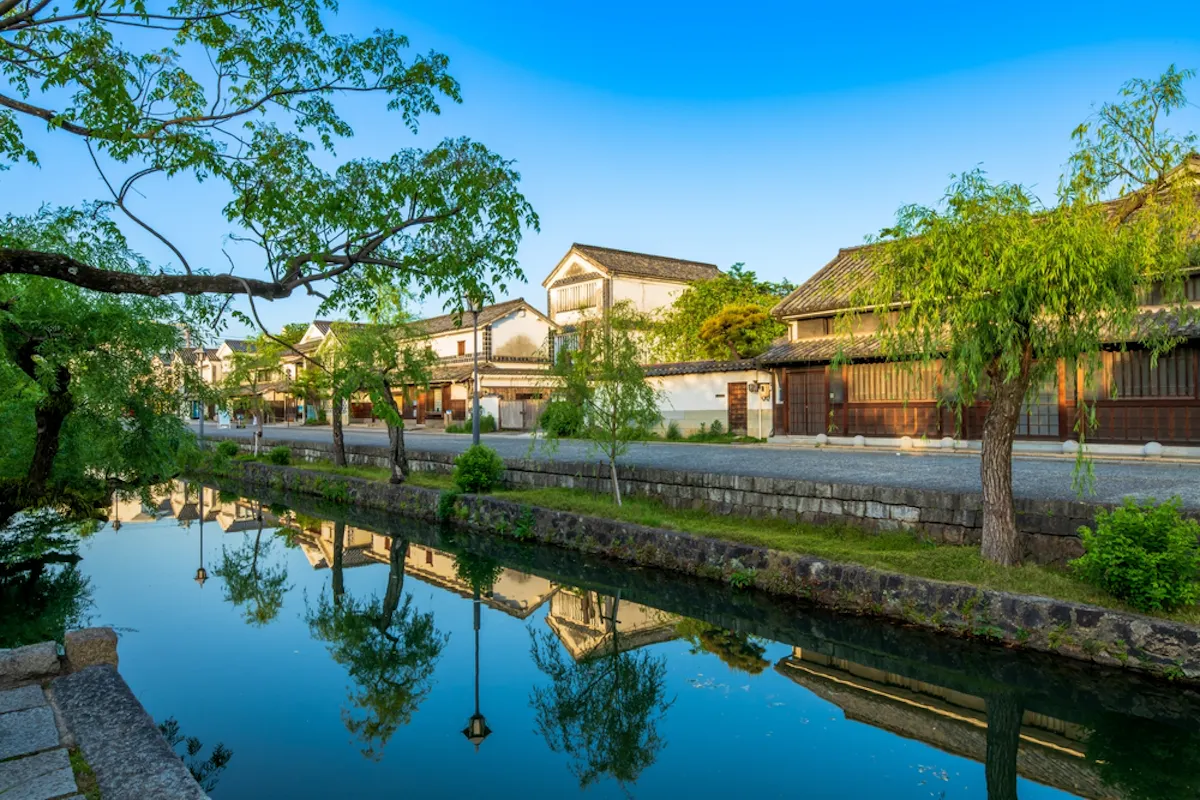 The old townscape of Kurashiki City, Okayama Prefecture A serene canal lined with lush green trees reflects traditional Japanese buildings under a clear blue sky. The scene is tranquil, with historic architecture and vibrant greenery mirrored in the still water, evoking a sense of peace and timeless beauty.