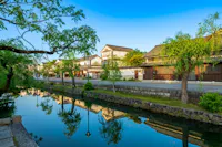 A serene canal lined with lush green trees reflects traditional Japanese buildings under a clear blue sky. The scene is tranquil, with historic architecture and vibrant greenery mirrored in the still water, evoking a sense of peace and timeless beauty.