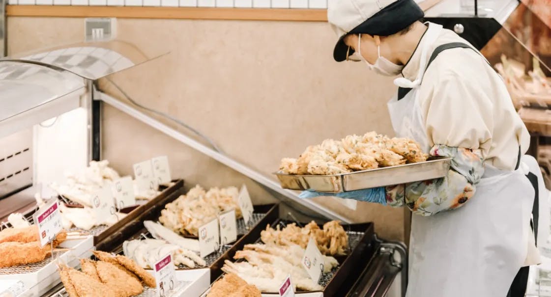 A person in a mask and apron arranges trays of fried food or tempura in a display case at a store or market.