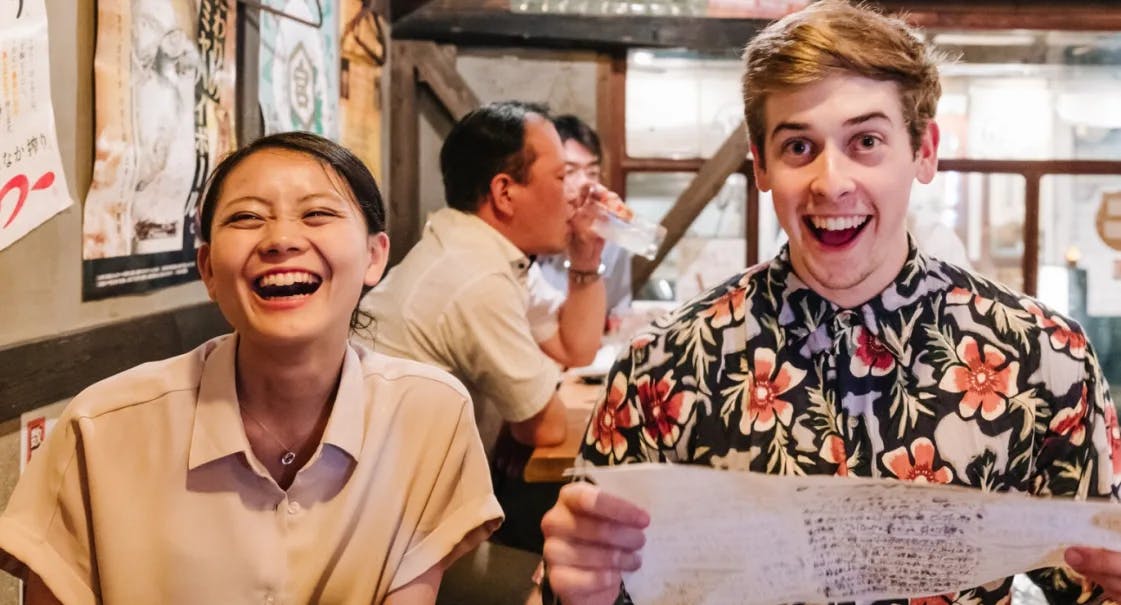 Two people sit side by side at a restaurant table, both smiling and laughing. The woman on the left wears a light blouse; the man on the right wears a floral shirt and holds a menu. Two other people are seated in the background.