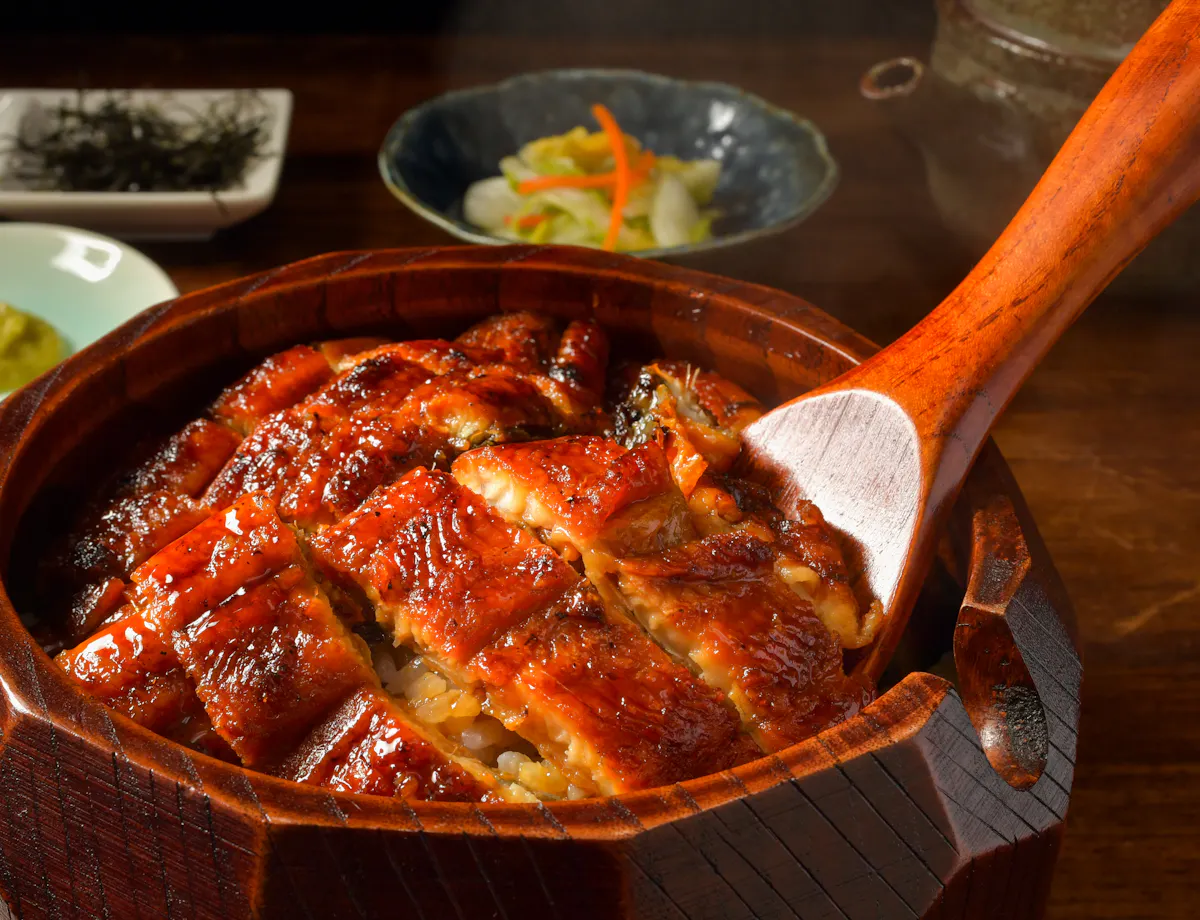 Hitsumabushi A wooden bowl filled with glazed grilled eel on rice, served with a wooden spoon. Small plates of pickles, wasabi, green onions, and seaweed are in the background on a wooden table.