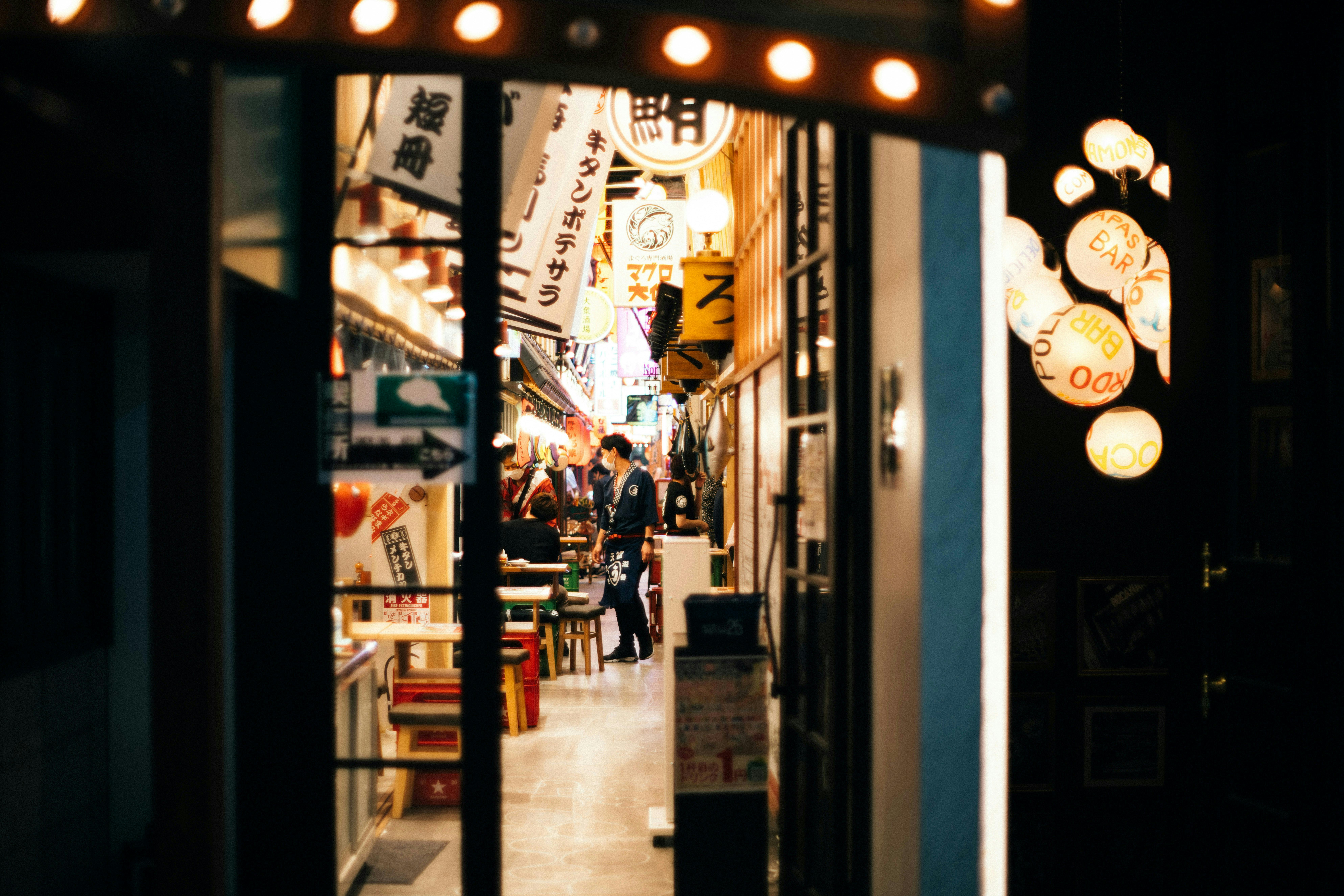 Narrow alleyway in Japan lined with lanterns and banners, leading to a small restaurant where people are gathered, with warm lighting creating a cozy atmosphere.