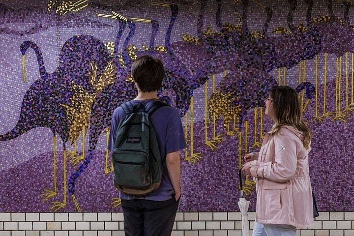 Two people stand facing a mosaic mural of ostriches with purple, blue, and gold tiles on a subway wall. The person on the left wears a backpack; the person on the right wears a pink jacket and holds an umbrella.