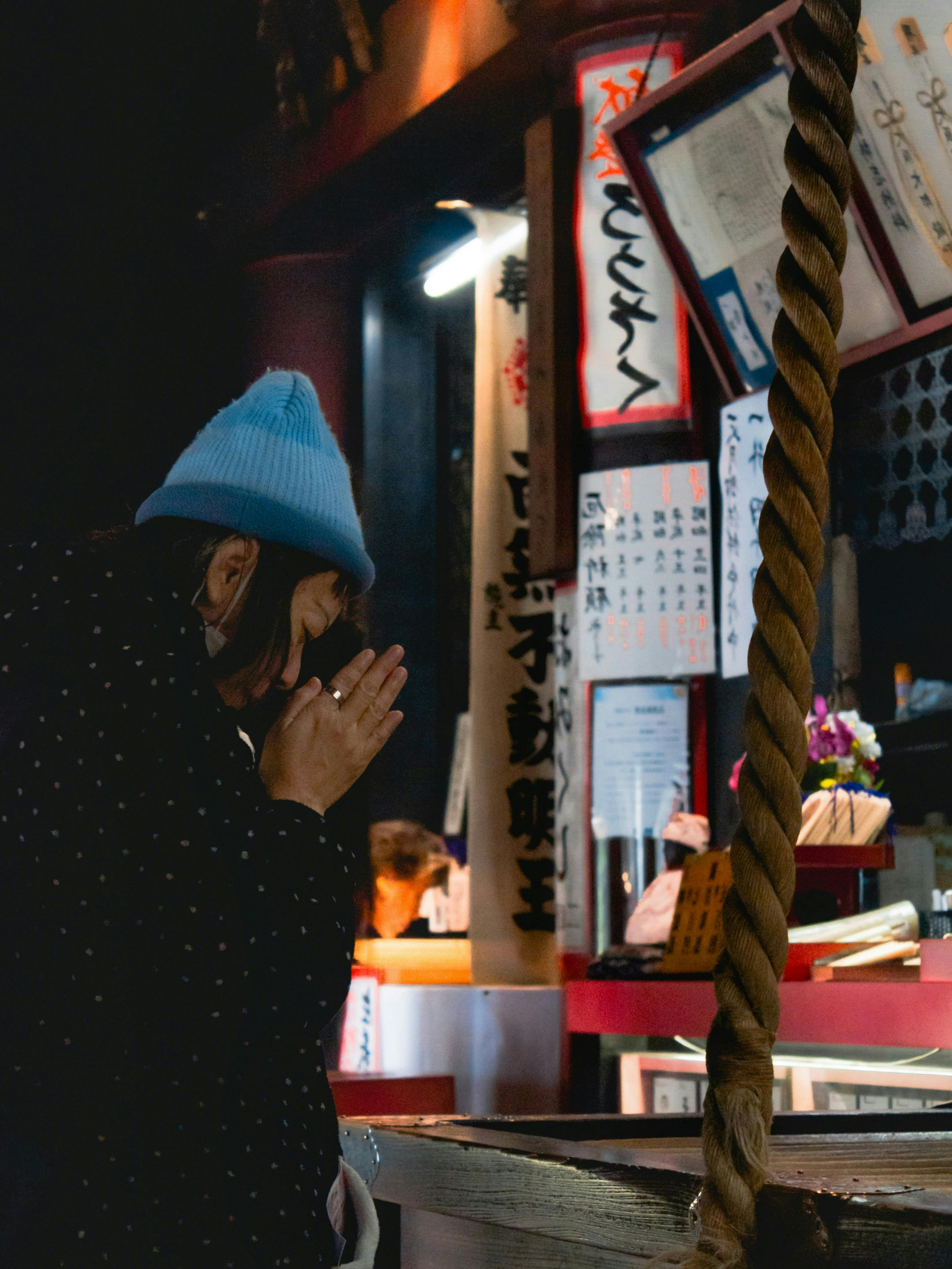A person wearing a light blue hat and dark coat stands in prayer with hands pressed together at a Japanese shrine, beside a thick rope and surrounded by traditional signs and offerings.