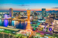 A vibrant cityscape of Kobe, Japan at dusk, featuring the illuminated Kobe Port Tower, a ferris wheel, modern buildings, and reflections on the waterfront under a colorful sky.