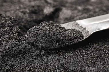 A metal scoop is resting in a mound of fine, black granular powder, with shining particles reflecting light on the surface.