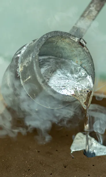 Sand Mold Casting A metal ladle pours hot, molten metal into a sand mold, with steam rising from the process. The scene depicts metal casting in a workshop environment.