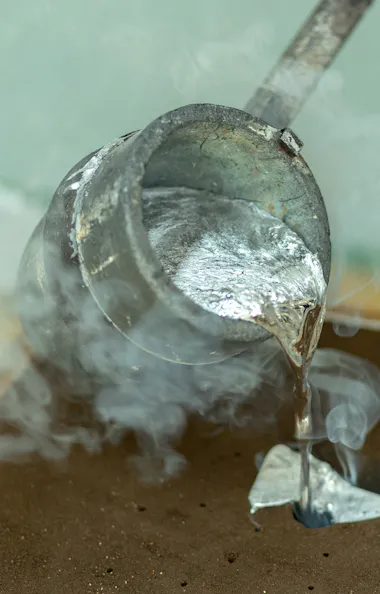 Sand Mold Casting A metal ladle pours hot, molten metal into a sand mold, with steam rising from the process. The scene depicts metal casting in a workshop environment.